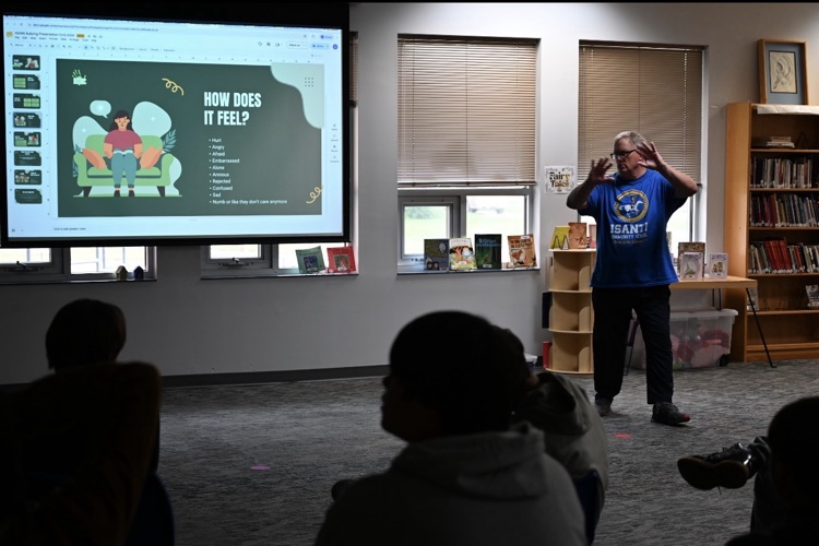 photo of a male teacher standing in front of slides projected onto the wall while the teacher is talking about bullying to students 