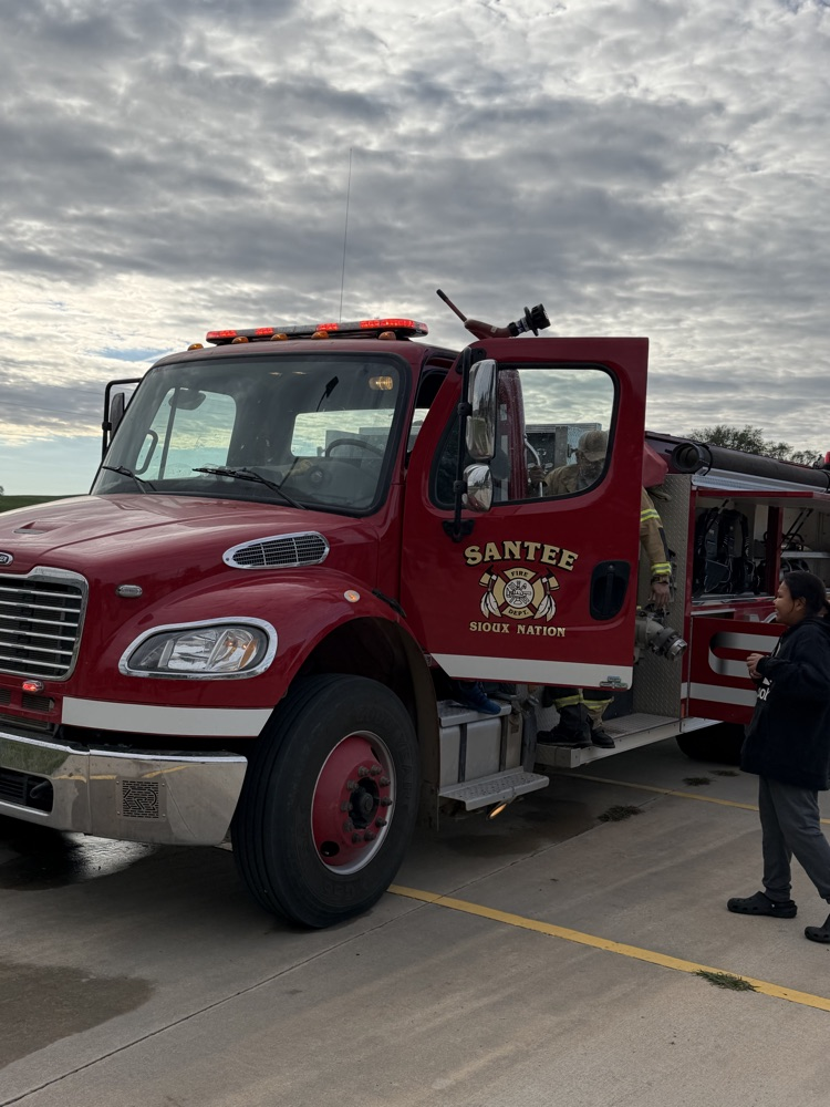 photo of a red fire truck and a male firefighter assisting a student onto the truck 