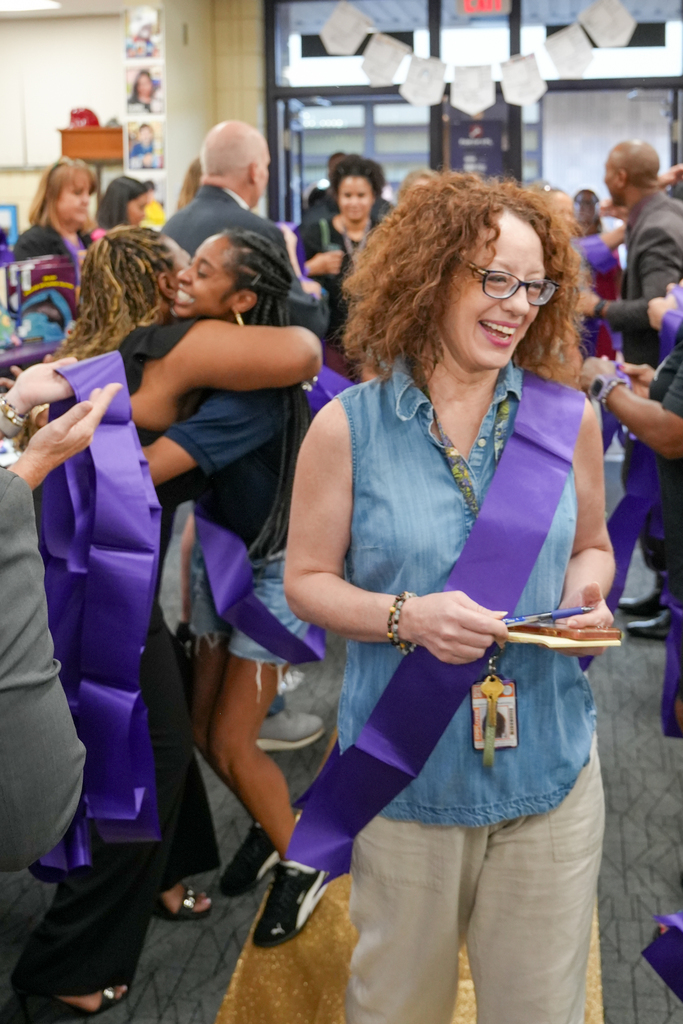 A woman with curly hair and glasses stands with a purple sash around her waist, smiling.