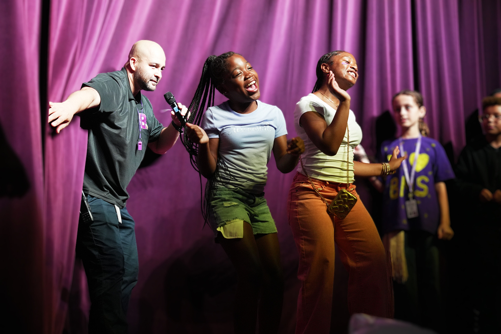 A man with a microphone stands behind two girls on stage. They all wear colorful outfits against a purple curtain.