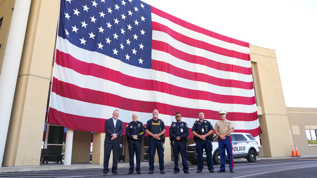 Police officers stand in front of a large American flag displayed on a building. A police vehicle is parked nearby.