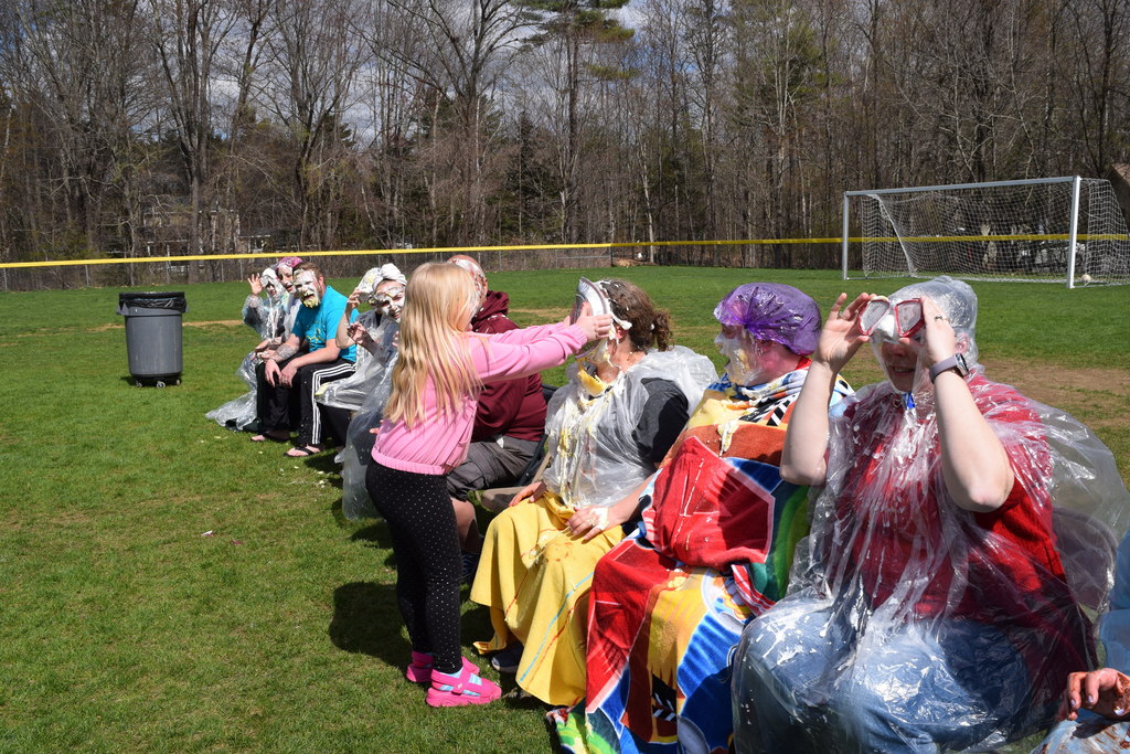 A student pieing a teacher in the face while other teachers look on in their ponchos and goggles.