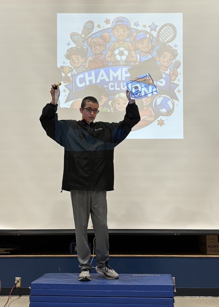 A student coach posing with his Champion's Club certificate on a podium made of stacked blue mats with the Champion's Club logo projected behind him.