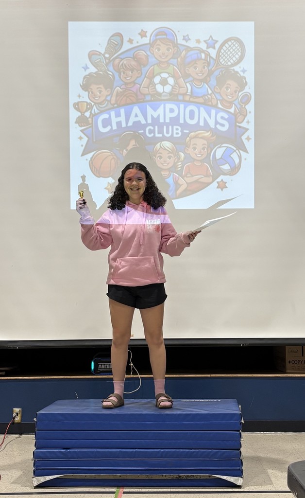 A student coach posing with her Champion's Club certificate on a podium made of stacked blue mats with the Champion's Club logo projected behind her.