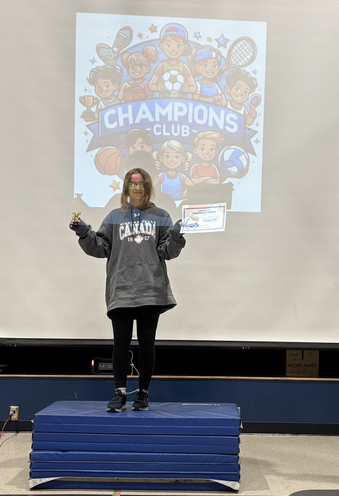 A student coach posing with her Champion's Club certificate on a podium made of stacked blue mats with the Champion's Club logo projected behind her.