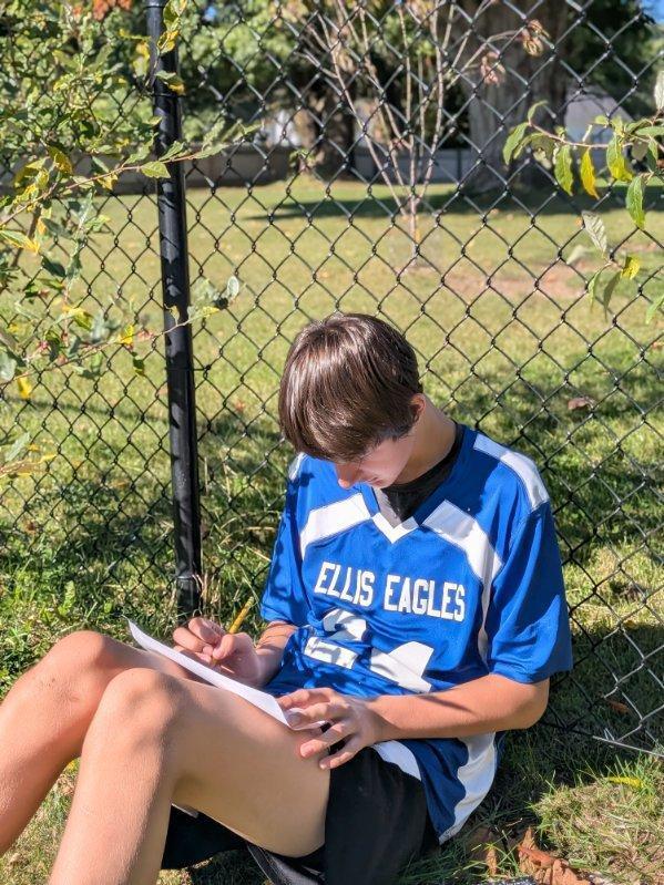 A student wearing an "Ellis Eagles" jersey sitting against the fence, writing in his notebook