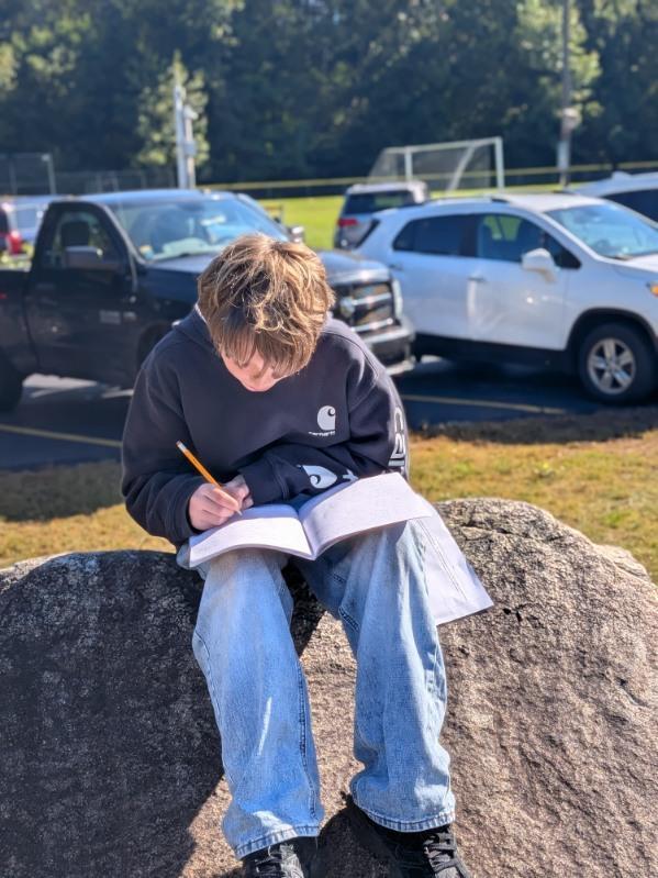 A student looking down while sitting on a rock, writing in his journal