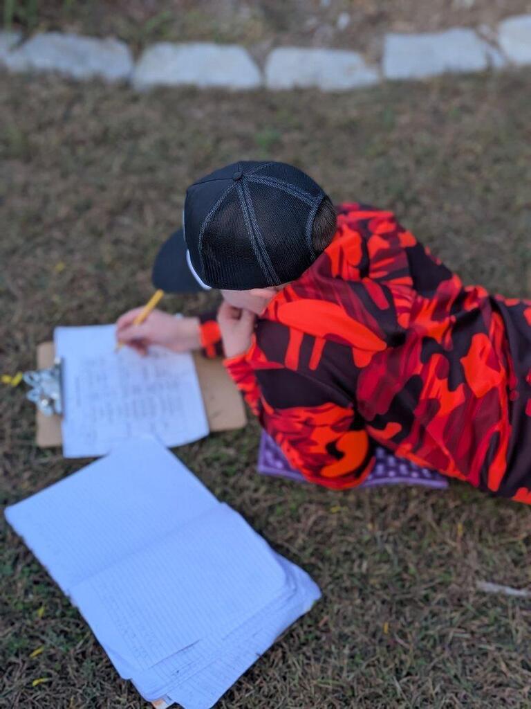 A student lying on the grass, filling out his notebook as well as a worksheet on a clipboard