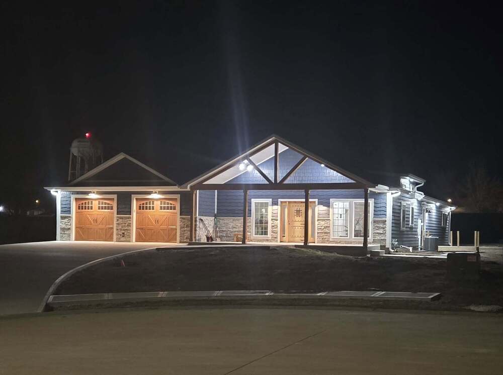 House light up by porchlights at night. The house features an attached 2 car garage, front porch, blue siding, and brick front.