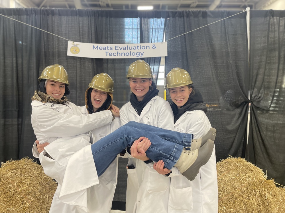 Three students pose for a picture while holding a 4th student across their arms. All students are wearing white lab coats and gold hard hats.