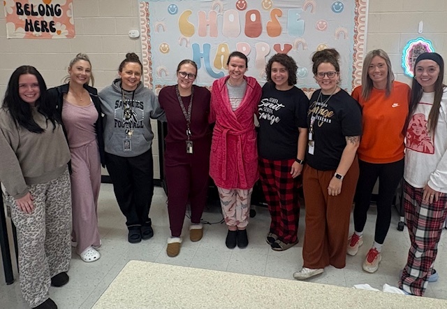 A group of eight women standing in a school hallway under a sign that says “Choose Happy.” They are wearing a variety of pajamas, robes, and slippers.