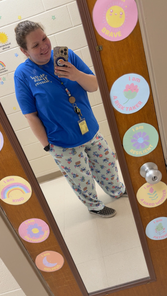 A woman taking a mirror selfie while smiling. She is wearing a bright blue school T-shirt and light pajama pants with colorful cartoon prints.