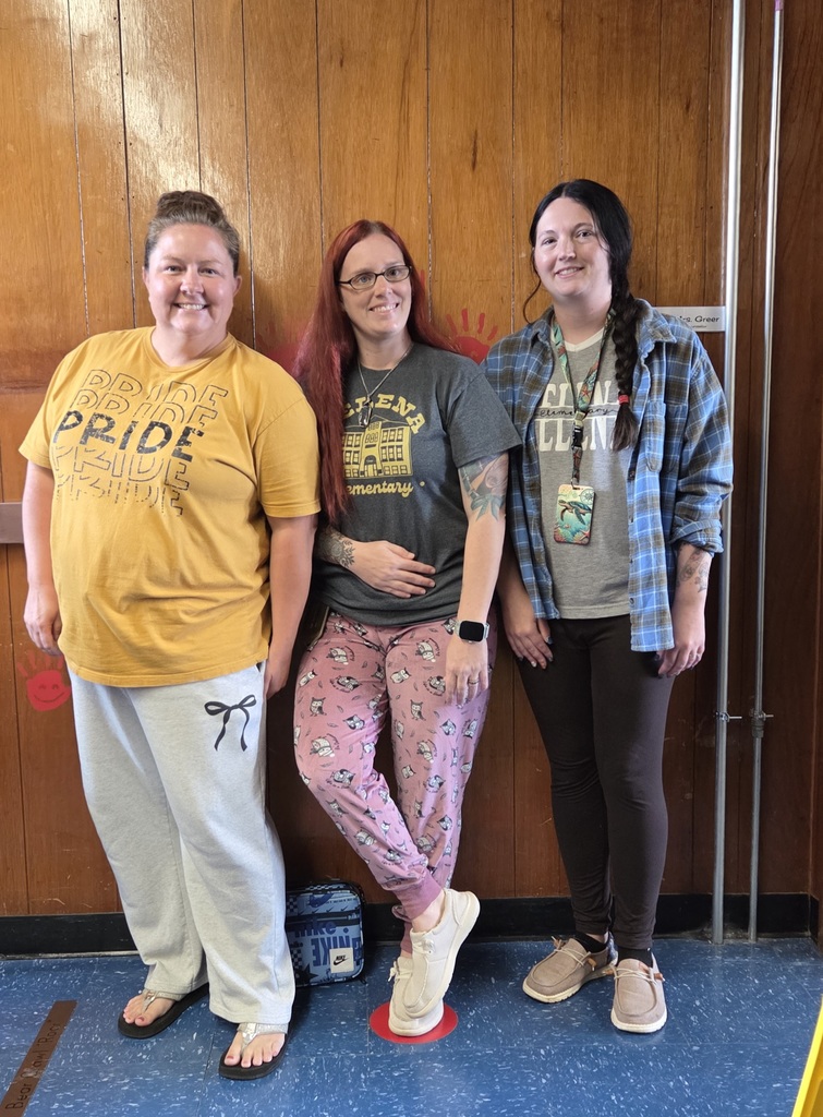 Three women smiling while standing against a wood-paneled wall. They are wearing casual pajama pants and school spirit shirts.