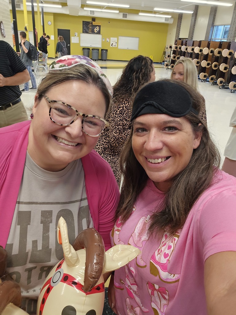 Two women smiling for a selfie in a school cafeteria. One wears a pink shirt with cartoon cowboy boots and a black sleep mask on her head, while the other wears glasses, a floral head wrap, and a gray shirt under a pink cardigan, holding an inflatable stick horse.