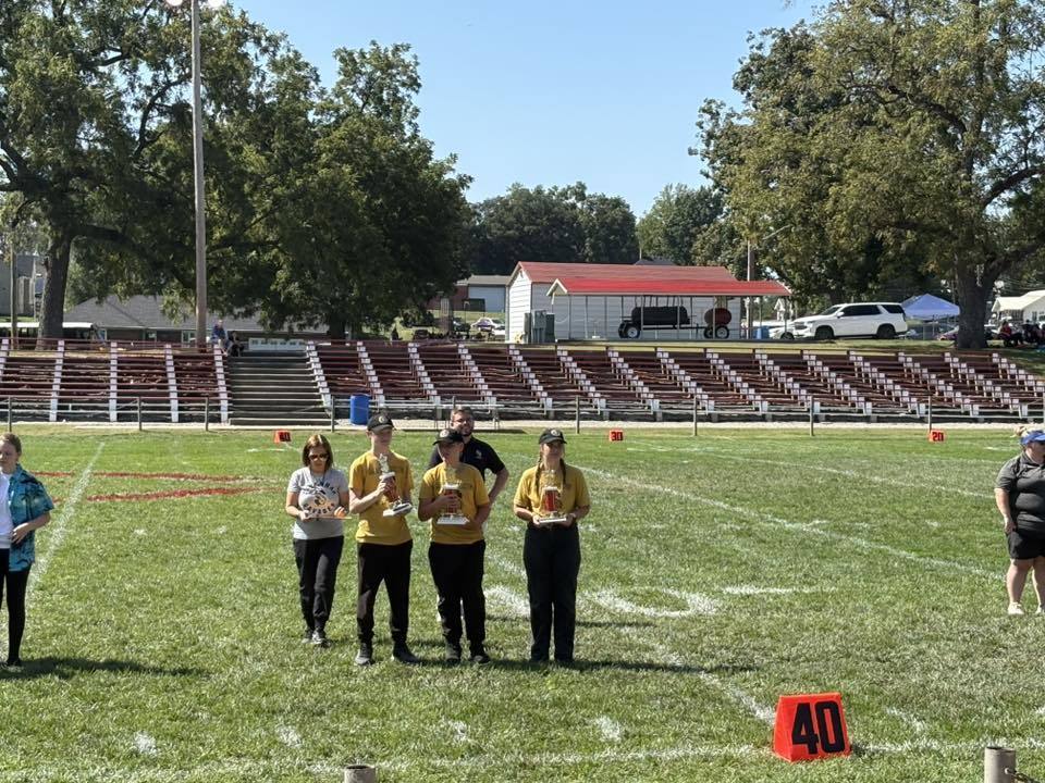 Three students stand on a football field holding trophies for the day. The band directors are behind them.