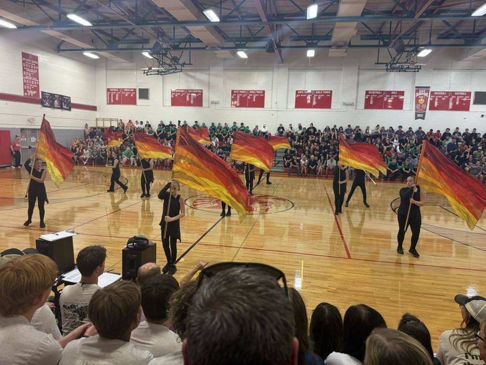 Several middle school color guard members are waving red, yellow, and gold flags as a packed gym watches.