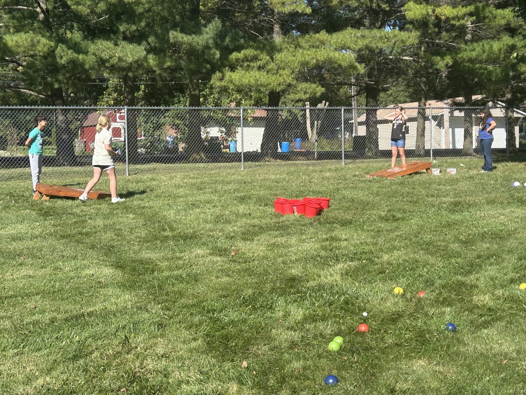 Student throws a beanbag toward a wooden cornhole board in a grassy yard.