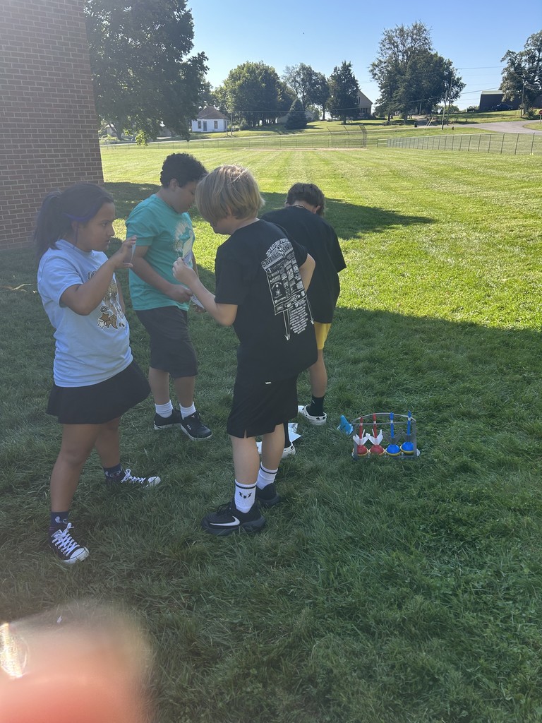 Group of four students talking near a brick wall, two holding papers while others listen.