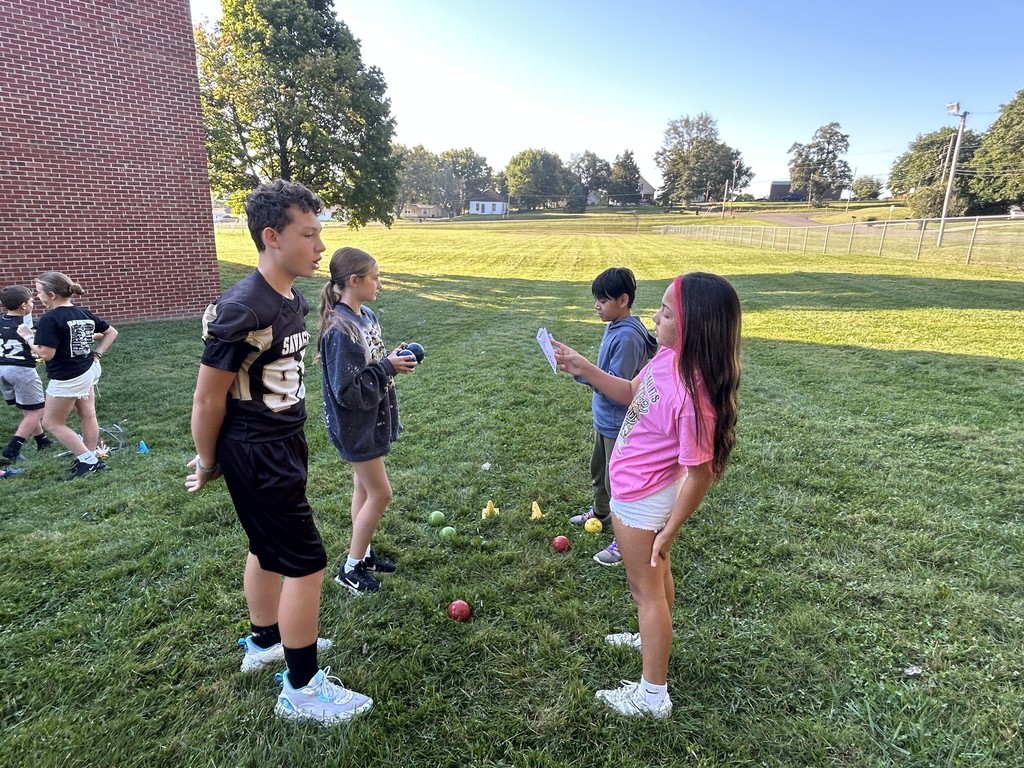 Four students stand in a circle on the grass, two holding papers and two holding game balls.