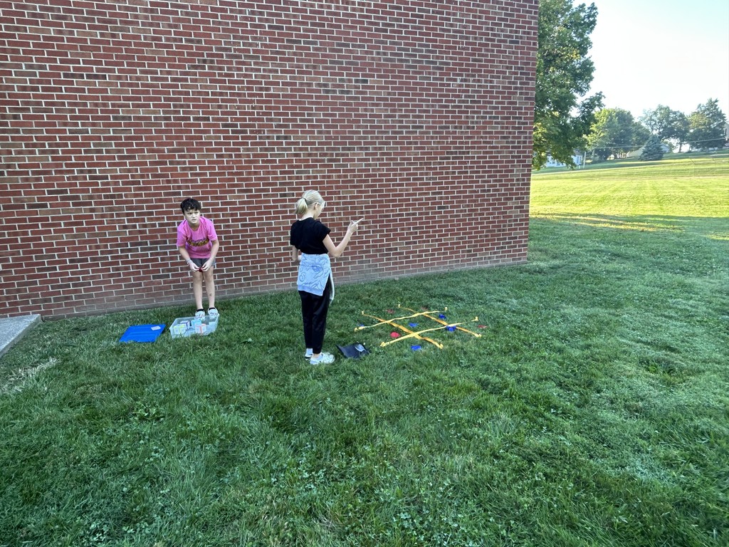 Two students play a tic-tac-toe toss game on the grass beside a brick wall.