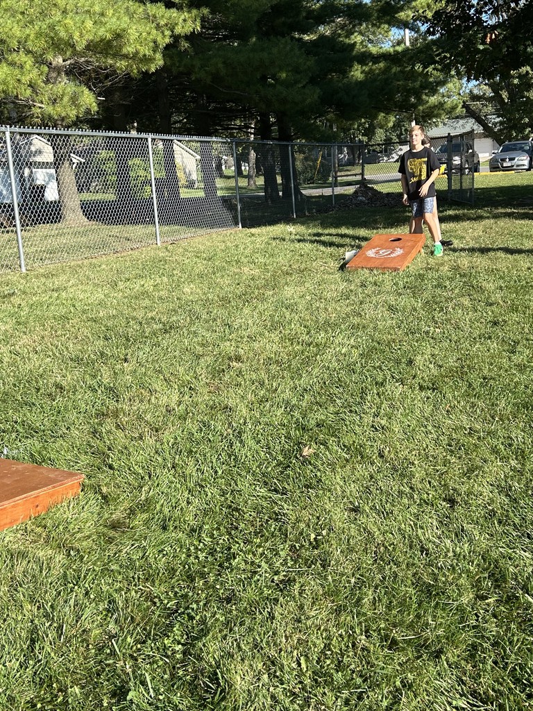 Student throws a beanbag toward a wooden cornhole board in a grassy yard.