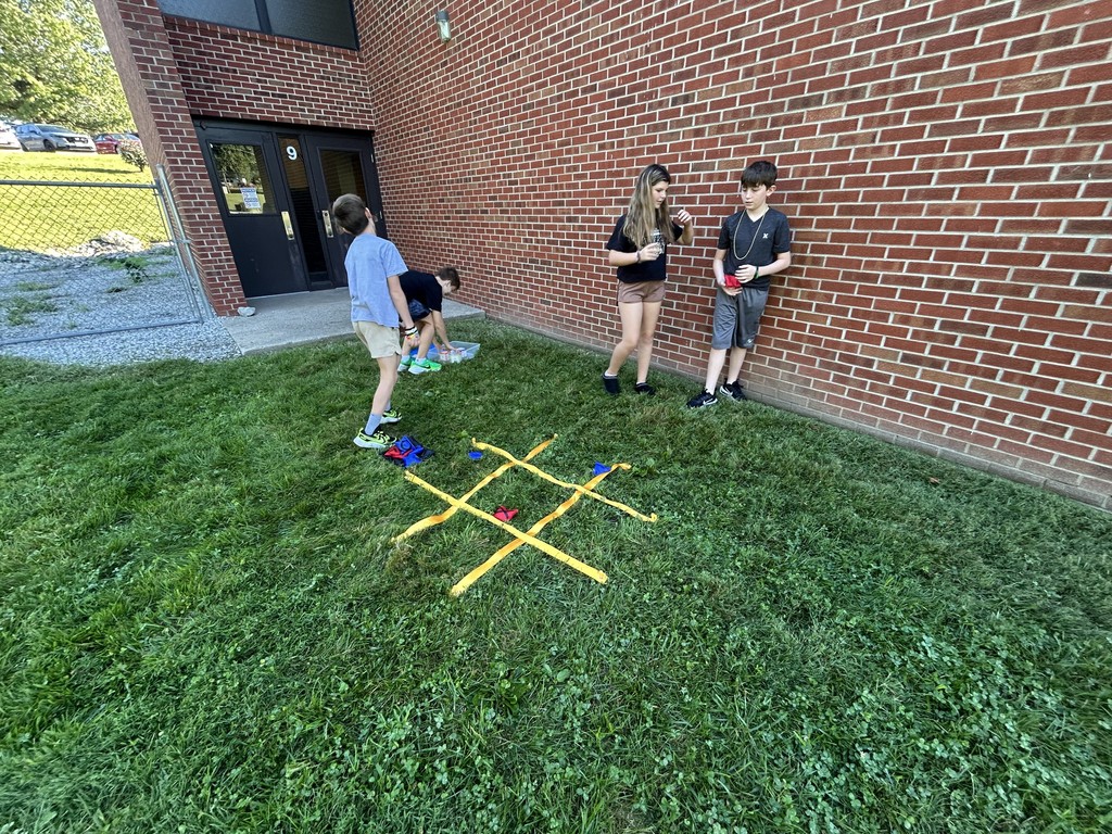 Four students play a beanbag tic-tac-toe game near a school building wall.