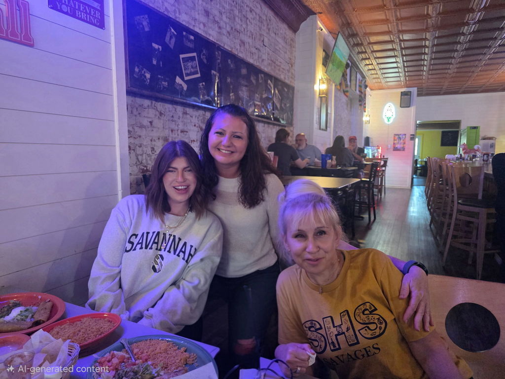 Three Savannah R-III staff members smile together at a restaurant table. One wears a light Savannah sweatshirt, one a mustard yellow SHS t-shirt, and one leaning in from behind is wearing a cream sweater. Plates of food are on the table in front of them.
