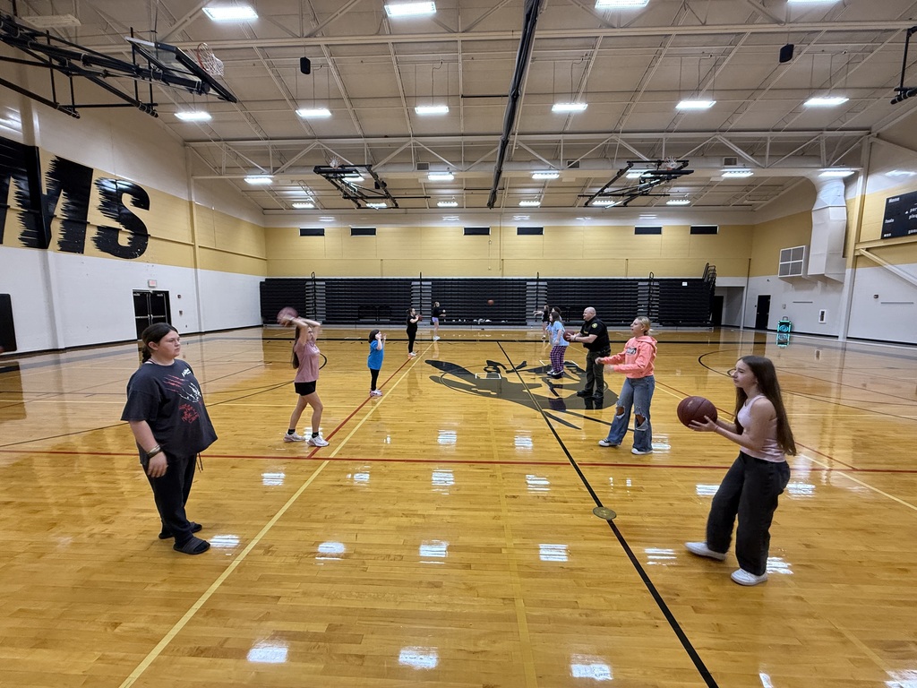 A school resource officer plays basketball with middle school students inside the gym, smiling and engaging during P.E. class.