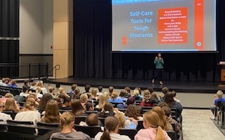 A guest speaker presents to high school students seated in the Performing Arts Center during a mental health awareness session.
