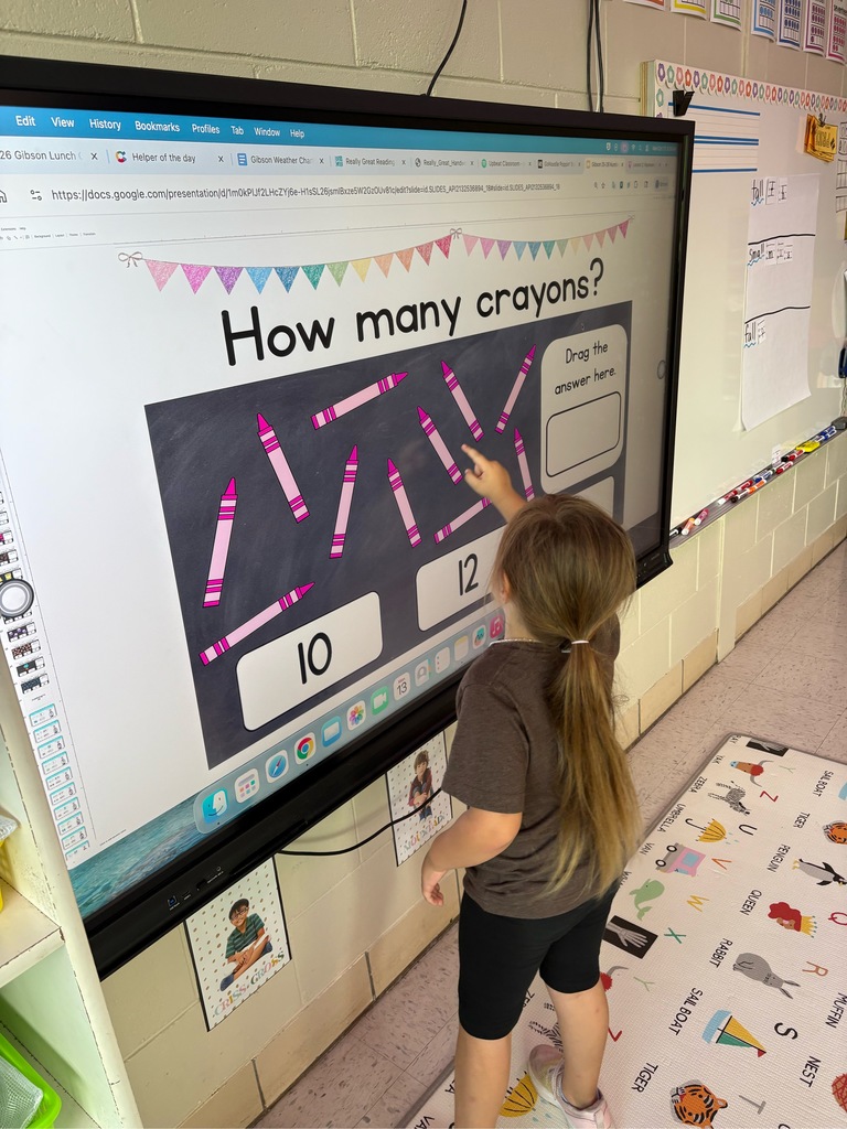 Large touch screen tv reads ‘How many crayons’. A young girl stands counting them. 