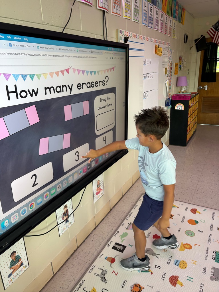 A large touch screen tv reads How Many Erasers.  A student stands in front of the screen and counts