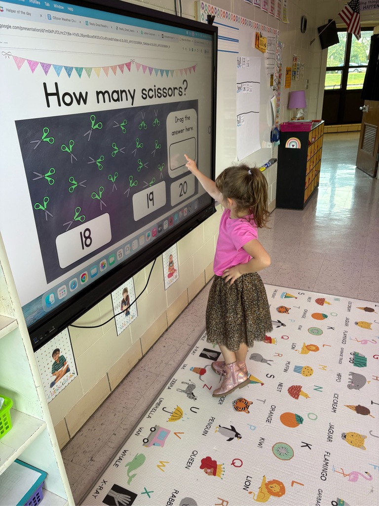 A large touch screen tv reads How Many Scissors, a young child stands in front of the screen and counts. 