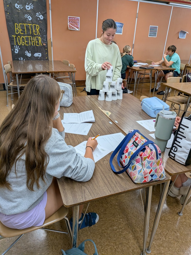students work in a classroom.  One student writes on paper while watching another student practice cup stacking.  students in the background are working on math projects. 