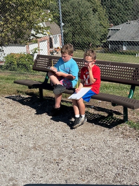 Two young boys sit together on a metal bench outdoors, writing on clipboards during a sunny day.