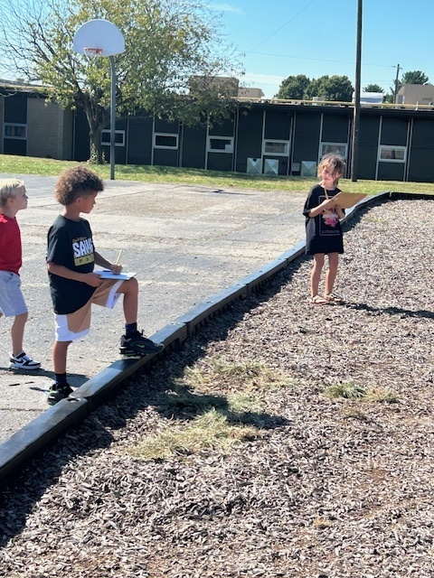 Three students stand along the playground border with clipboards, drawing or writing.