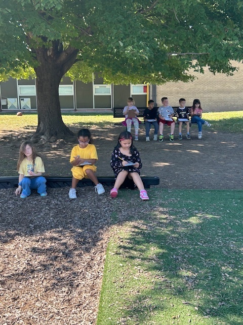 A group of elementary students sit in the shade under a large tree, some on benches and others on the ground, working on assignments.