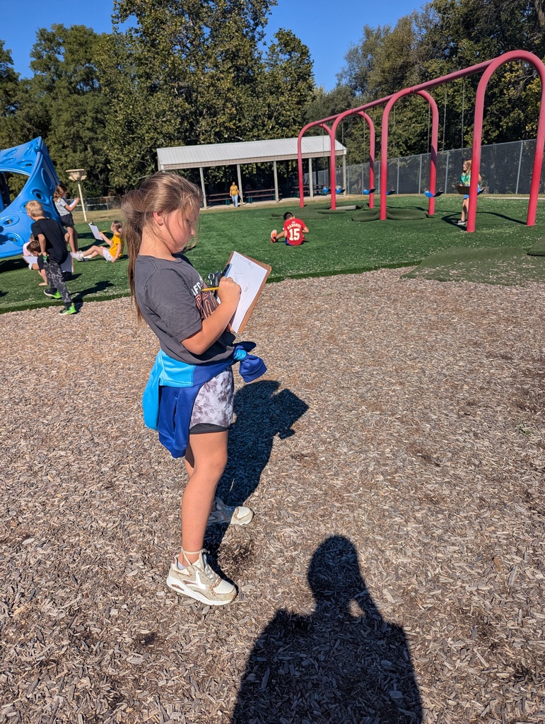 A young girl stands on the playground holding a clipboard and pencil, sketching while classmates play in the background near swings.