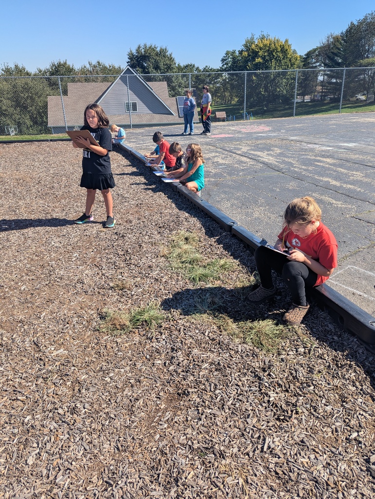 Several students kneel or sit along the playground border with clipboards, drawing or writing while others walk nearby on the blacktop.