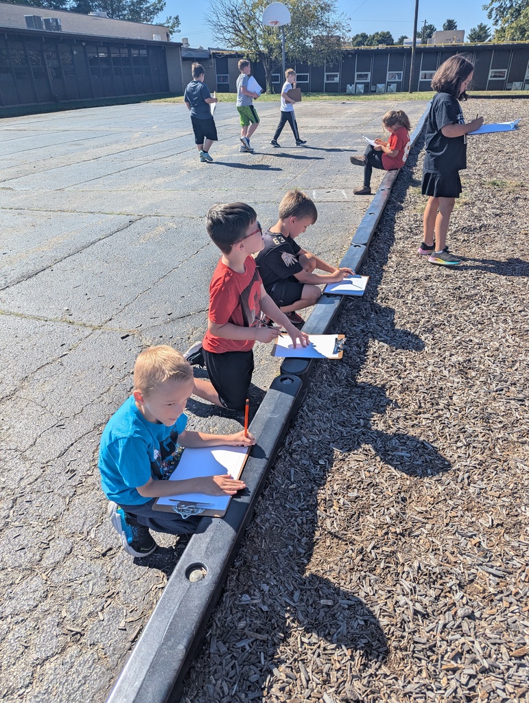 Several students kneel or sit along the playground border with clipboards, drawing or writing while others walk nearby on the blacktop.