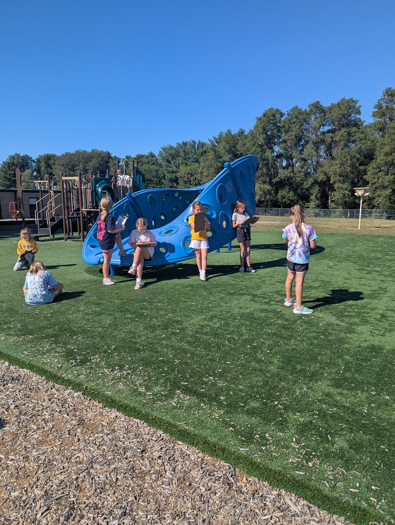 A group of students sit and stand around a large blue climbing structure on the playground, drawing on clipboards under a clear blue sky.