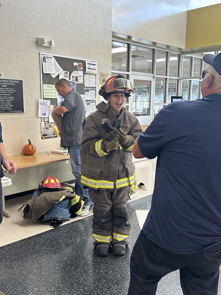 A high school student in firefighter turnout gear laughs during a Gear Challenge activity as a firefighter records the moment on a phone.