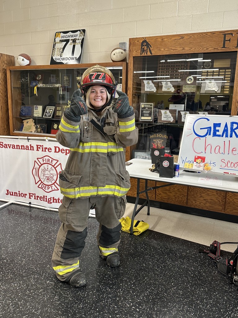 A high school student in full firefighter gear gives two thumbs up while standing near a Savannah Fire Department Junior Firefighter display table.
