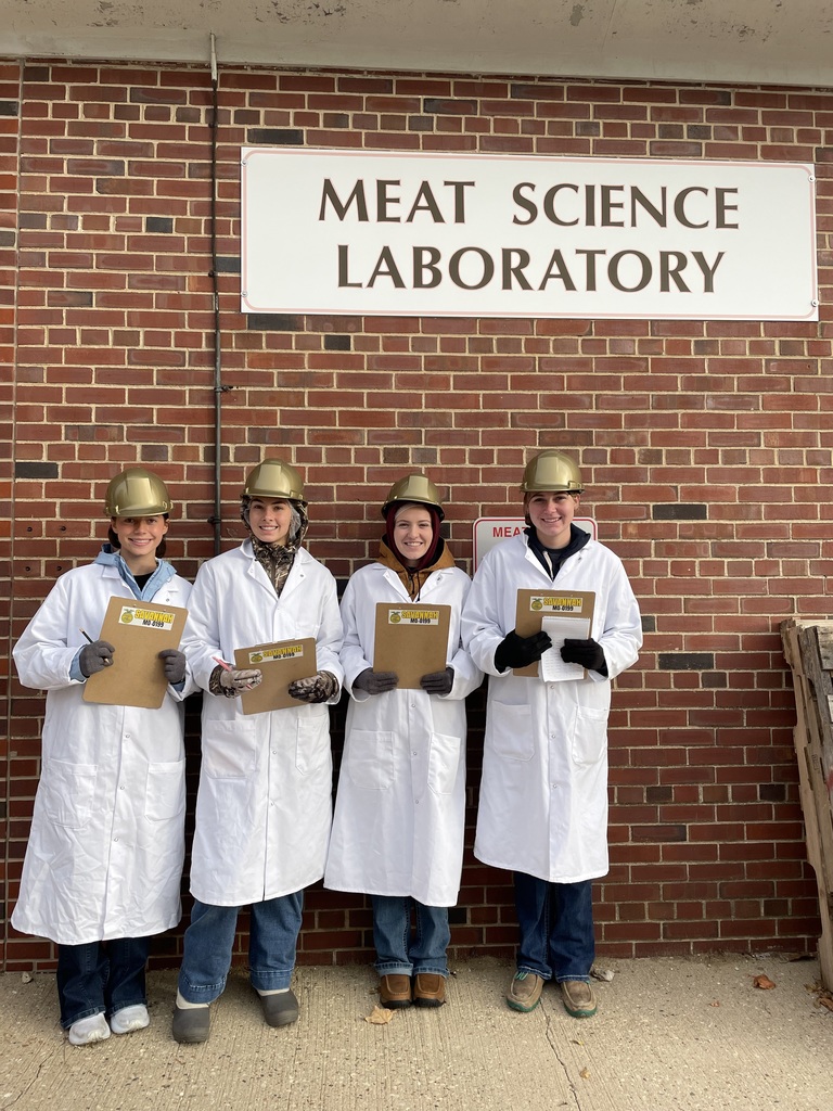 group of students in white lab coats and gold hard hats hold clip boards and pose against a brick will under a sign that reads 'Meat Science Laboratory" 