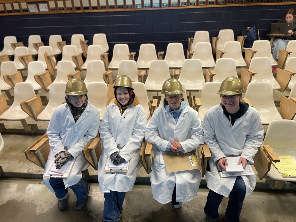 Four students in lab coats and helmets sit with clipboards in tiered classroom seating, smiling.