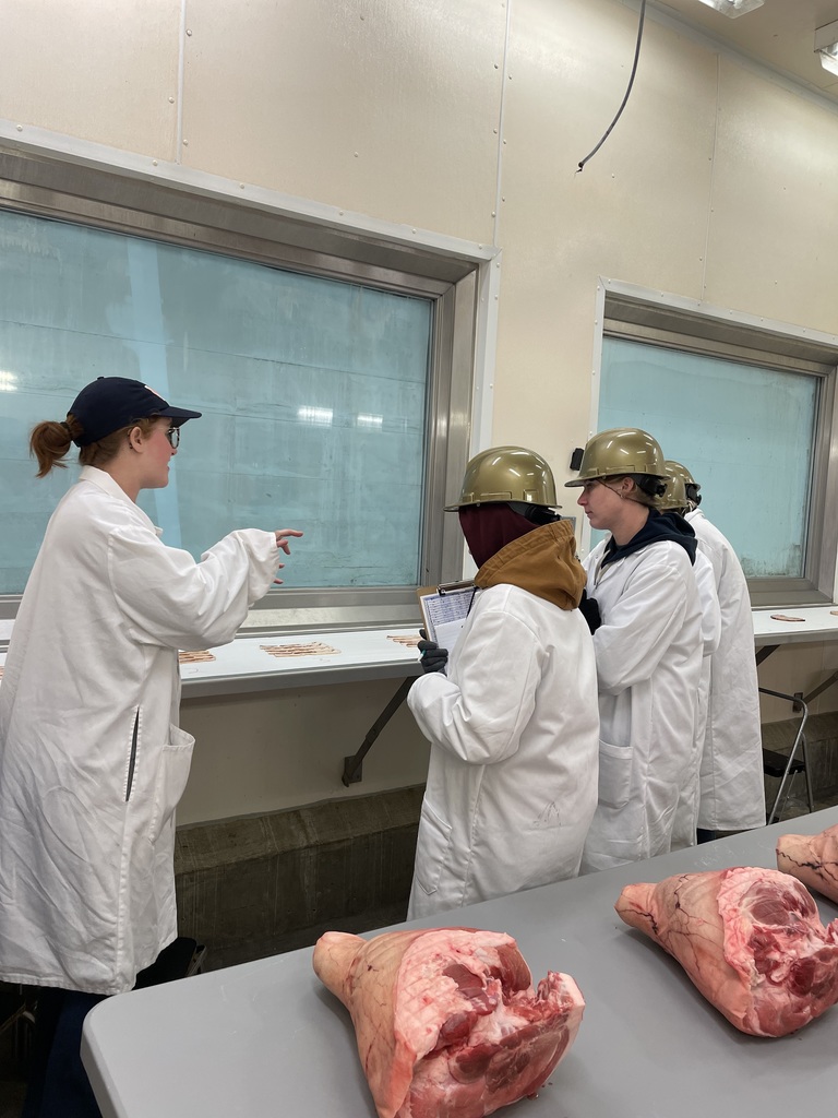 Instructor gestures toward sliced meat samples as students in lab coats and helmets take notes.