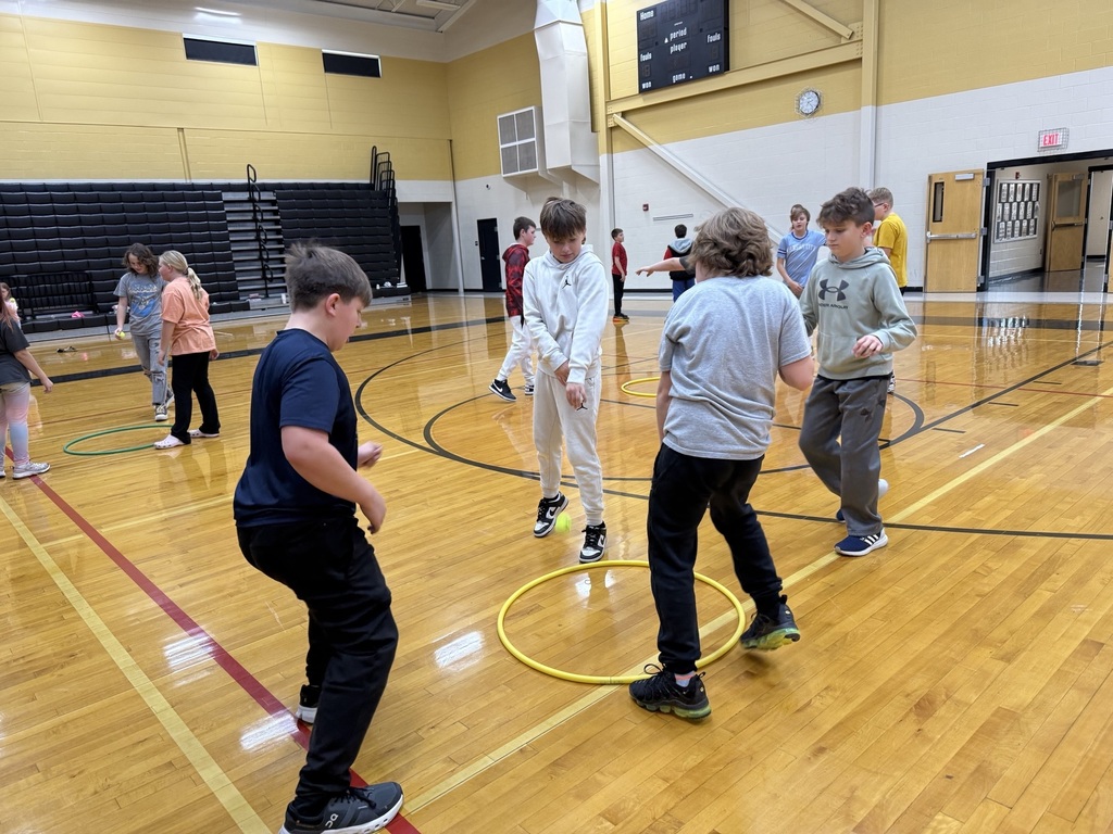 Students gather around hula hoops on the gym floor, focusing as they play a Spikeball-style game during PE class.