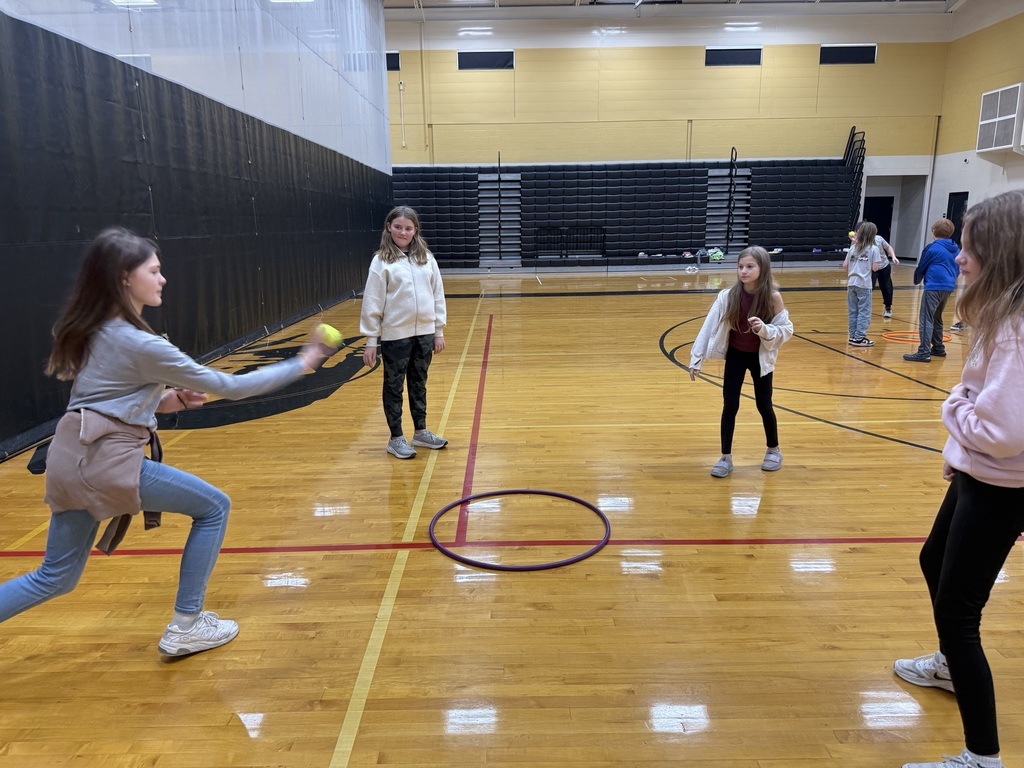 Four girls stand in a gym tossing a yellow ball while playing Spikeball, smiling and cheering each other on.