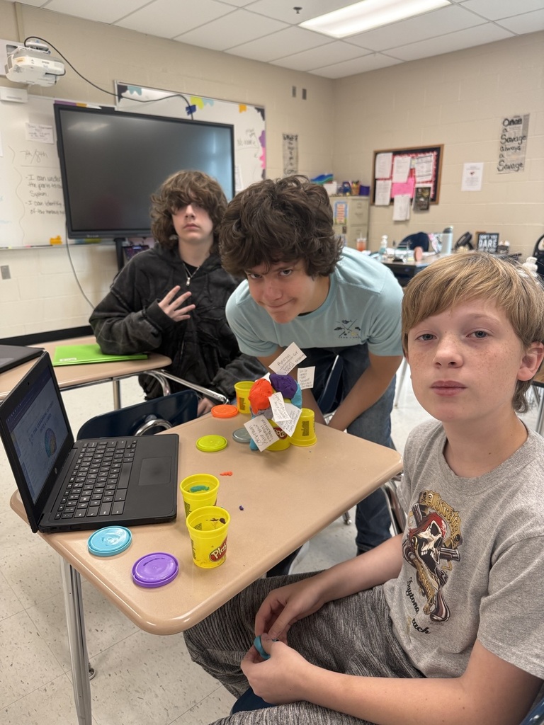Three students sit at a classroom desk using colorful Play-Doh to build a brain model beside a laptop.