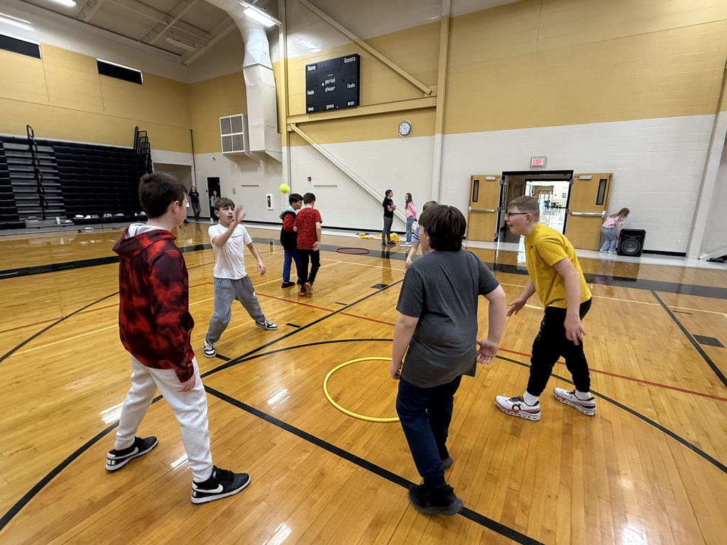 A group of middle school boys play Spikeball inside the gym, laughing as one tosses a ball toward the others.