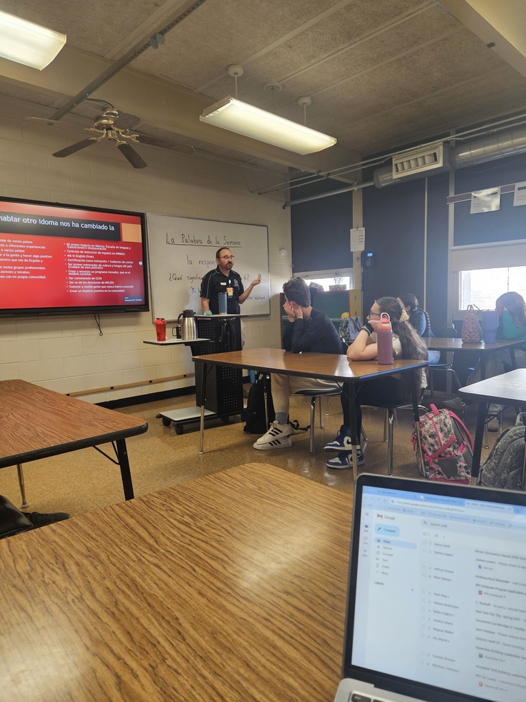 A speaker stands in front of the classroom gesturing toward a slide in Spanish about how speaking another language has changed their life.